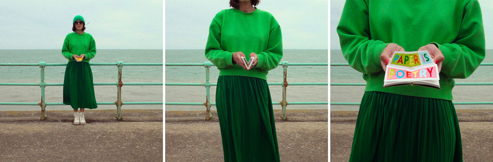 Person in gr&uuml;ner Kleidung steht an einer Strandpromenade vor dem Meer und h&auml;lt ein kleines aufgeklapptes Buch mit der Aufschrift Paper is Poetry in den H&auml;nden